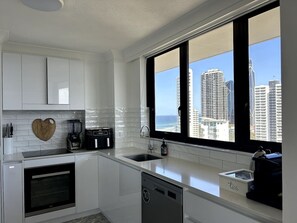 A modern kitchen with white cabinets, a heart-shaped cutting board, a coffee maker, and a view of tall buildings through large windows. - Beachside Escape, VIEWS all around, steps to beach (Surfers Paradise)