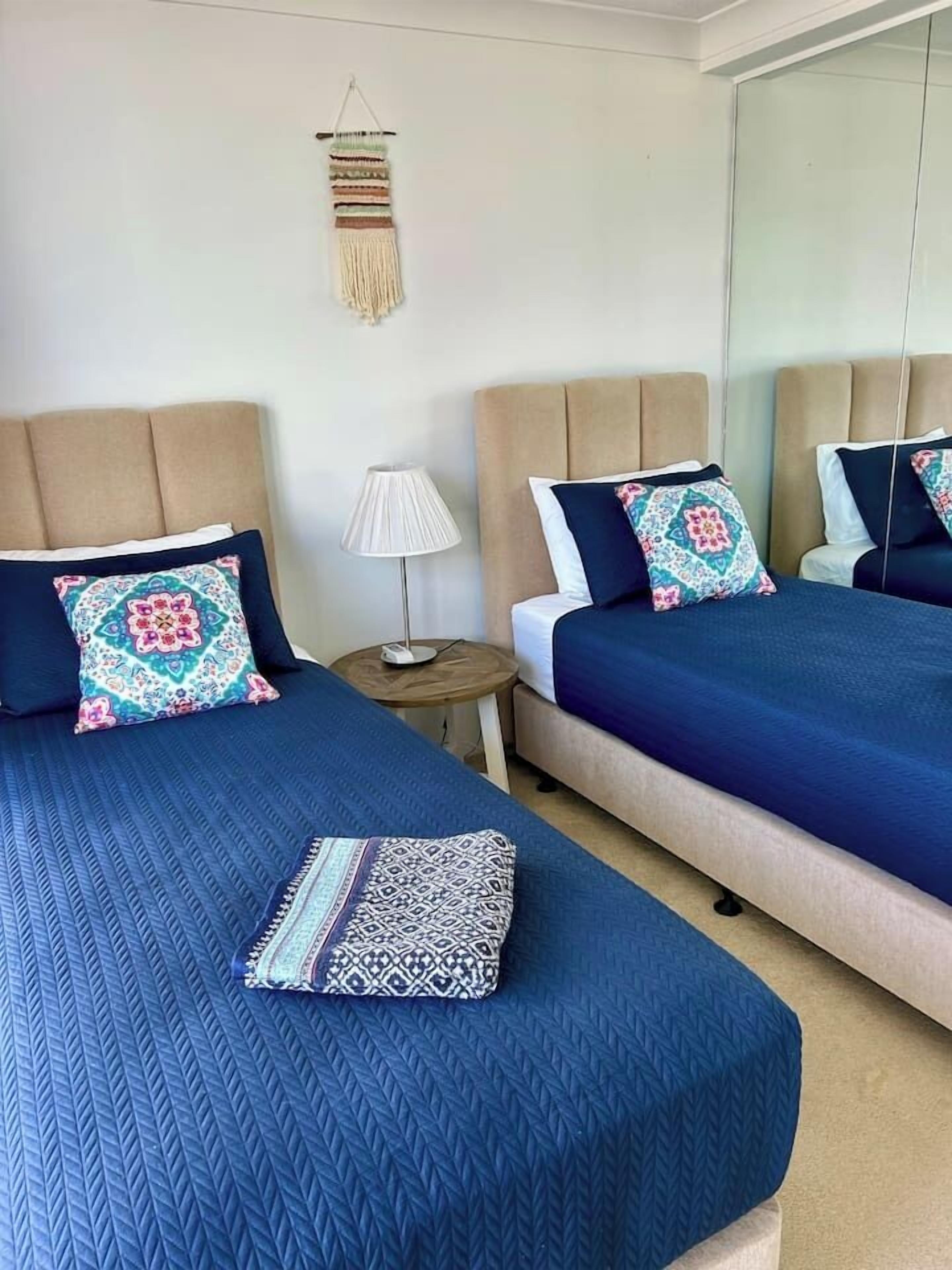 Une chambre d’hôtel avec deux lits, une table de chevet avec une lampe et une décoration murale en macramé.