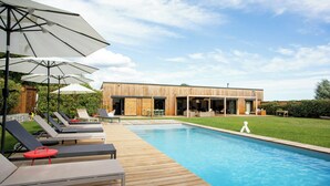 Une terrasse en bois avec des chaises longues et des parasols, une piscine et un bâtiment moderne doté de grandes fenêtres.