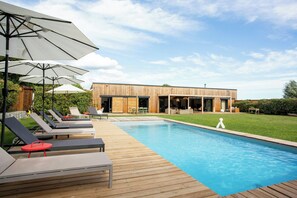 Eine Holzterrasse mit Liegestühlen und Sonnenschirmen, ein Schwimmbecken und ein modernes Gebäude mit großen Fenstern.