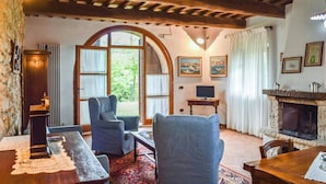 Ein gemütliches Zimmer mit Holzbalkendecke, Kamin, Fernseher und einem großen Fenster mit Vorhängen.