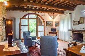 Ein gemütliches Zimmer mit Holzbalkendecke, Kamin, Fernseher und einem großen Fenster mit Vorhängen.