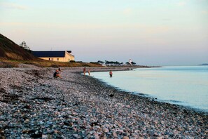 A pebbled beach with a building and a lighthouse in the distance. - Minimalist cottage with stunning sea view (Knebel)