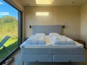 A modern bedroom with a large bed, a view of greenery outside, and a skylight above. - Minimalist cottage with stunning sea view (Knebel)