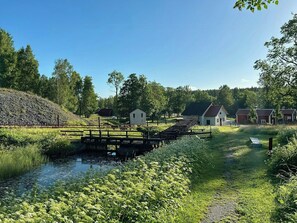 Ländliche Landschaft mit einer kleinen Brücke über einem Bach, umgeben von viel Grün und einigen Gebäuden.