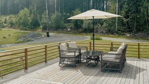 Terraza con mobiliario de mimbre y una mesa, rodeada por una barandilla de madera, con vistas a un área boscosa.