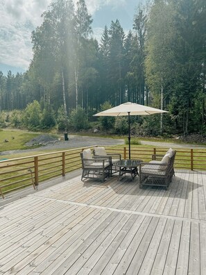 Eine Terrasse mit Korbmöbeln und einem Tisch, umgeben von einem Holgitter, mit Blick auf einen bewaldeten Bereich.