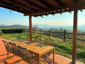 An outdoor wooden deck with a table and chairs, overlooking a grassy landscape with distant hills. - Tuscan apartment with panoramic views in Iano (Montaione FI)