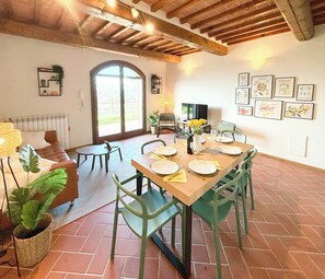 A dining area with a wooden table set for four, green chairs, a television, and a large window with a view of greenery. - Tuscan apartment with panoramic views in Iano (Montaione FI)