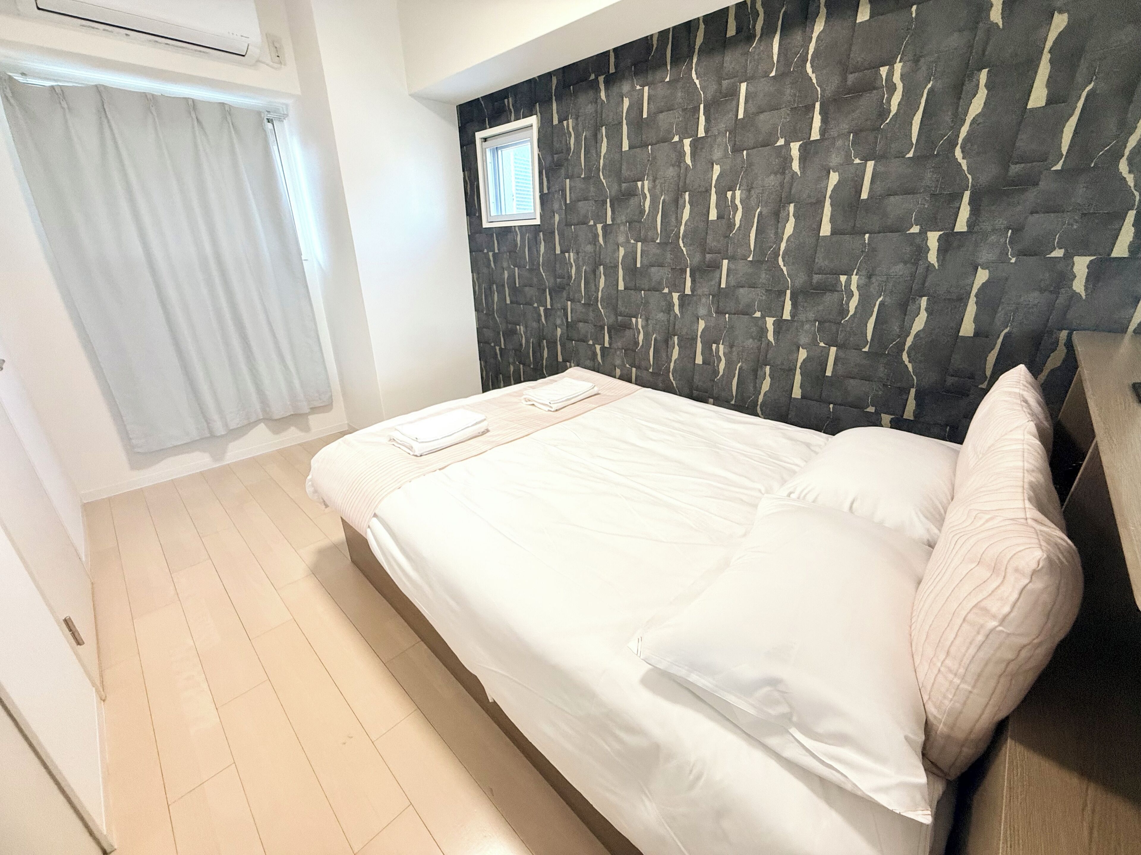 A hotel room with a bed, a window with curtains, a stone-patterned headboard, and a small window above the bed.