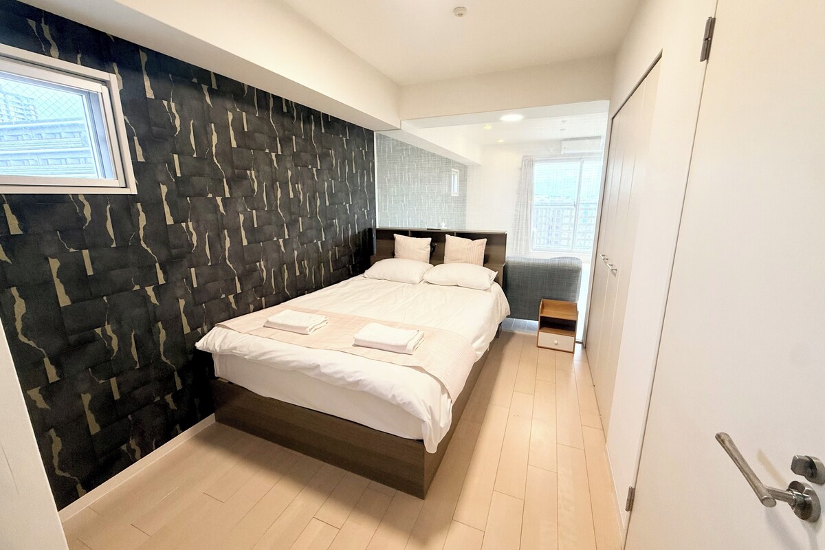A modern hotel room with a large bed, a dark patterned wall, a small bedside table, and a view of the city through a window.