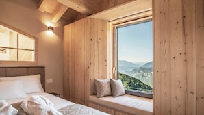 A wooden-paneled room with a bed, a window seat with pillows, and a view of a mountainous landscape. - Alpine flats in Fai della Paganella (Fai della Paganella)