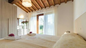 A bedroom with a large bed, a wooden ceiling, a window with curtains, a bathroom with a mirror, and a vase with flowers. - Orlando. Villa with private pool near Lucca (Colle di Compito)