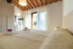 Ein Schlafzimmer mit einem großen Bett, Holzboden, einem Fenster mit Vorhängen, einem Badezimmer mit Spiegel und einer Vase mit Blumen.