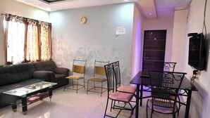 Una sala de estar con un sofá, una mesa de centro de vidrio, una mesa de comedor con sillas y un televisor montado en la pared.