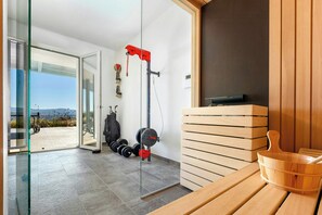 A modern gym with weightlifting equipment, a sauna, and a view through a glass door. - Exclusive villa with panoramic pool and gym (Campagnatico)