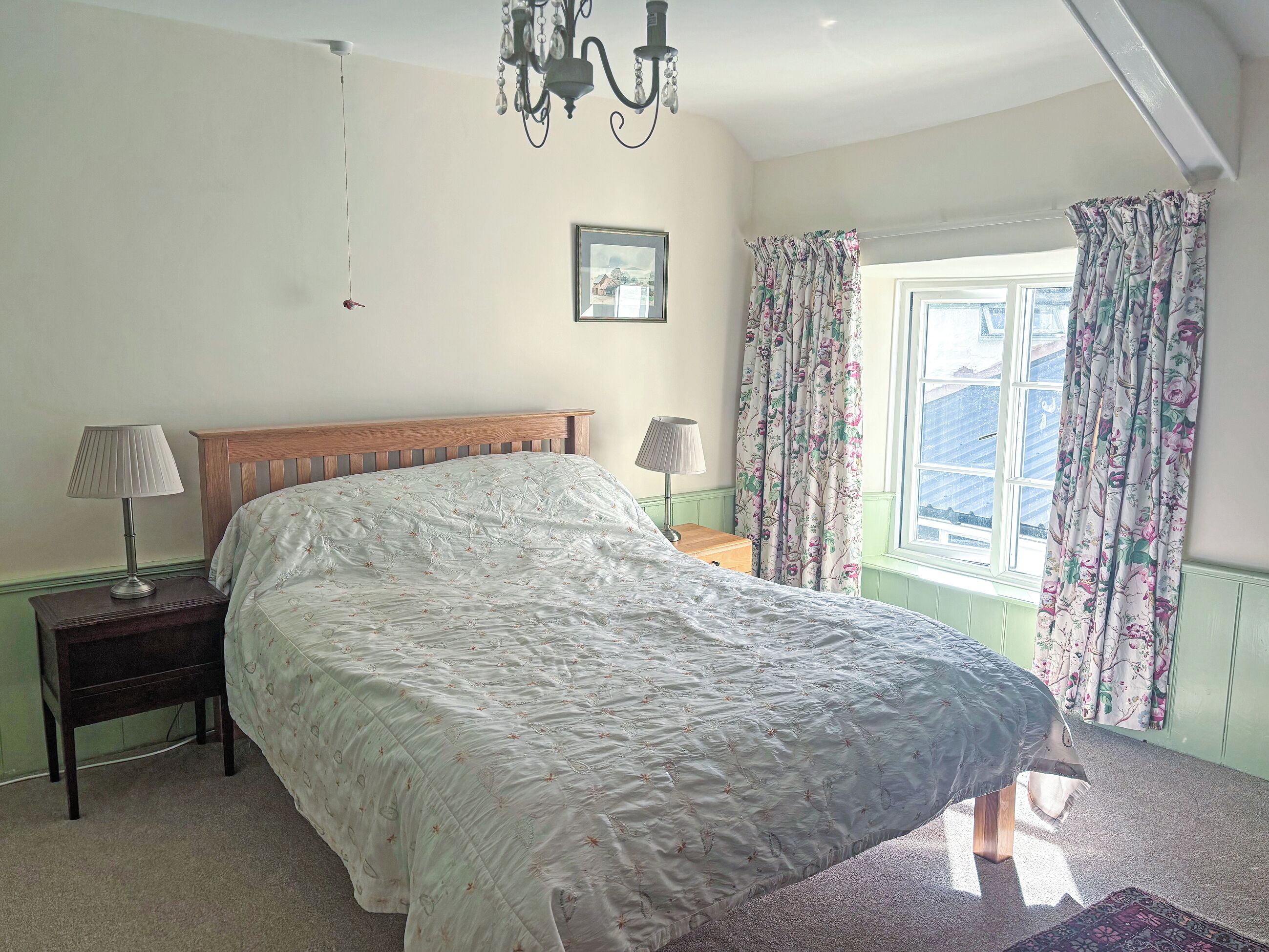 A bedroom with a large bed, a wooden headboard, a nightstand with a lamp, a picture on the wall, and a window with floral curtains.