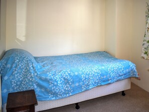 Uma cama de solteiro com colcha azul florida em quarto com paredes brancas e uma pequena mesa de cabeceira de madeira.