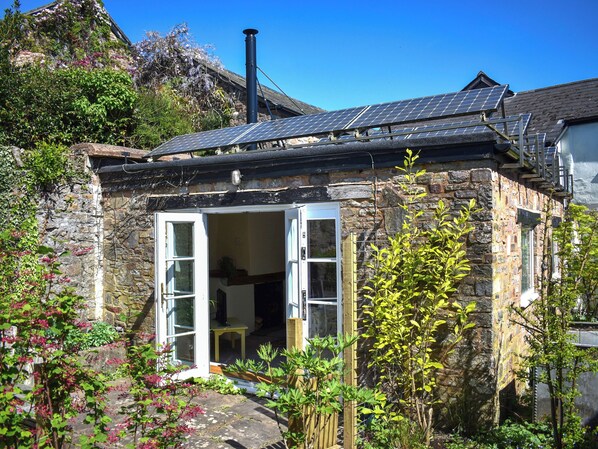 Ein Steinhäuschen mit Solardach, Schornstein und einem Garten mit blühenden Pflanzen.