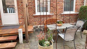 En murstensbygning med en trædør, en metalpergola, udendørs siddepladser og en fletkurv med blomster.