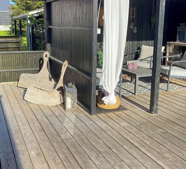 Eine Holzterrasse mit einem Korbstuhl, einem kleinen Tisch und einem weißen Vorhang.