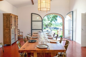 A dining area with a long wooden table set for six, surrounded by chairs, with a view through an arched window. - Historic Provençal farmhouse with pool in Provence (Le Plan-de-la-Tour)
