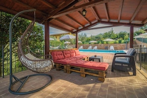 Ein überdachter Außenbereich mit Hängestuhl, einer roten gepolsterten Couch und einem Pool im Hintergrund.