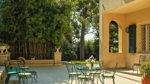 Une terrasse extérieure avec une table et des chaises, entourée de verdure et bordée d’un bâtiment doté d’une arche.