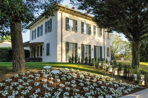 Un edificio a due piani con facciata simmetrica, un portico coperto e un giardino curato con fiori bianchi.