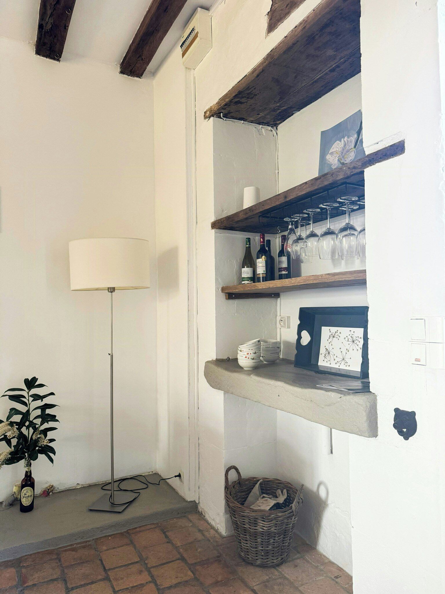 A room with a lamp, shelves with bottles and glasses, a basket, and a plant.