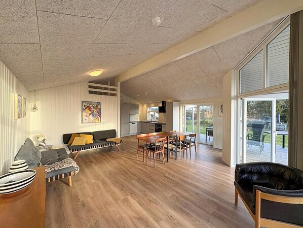 A spacious living area with a sofa, dining table, and chairs, a kitchen in the background, and large windows. - Holiday home close to beach near Norsminde (Odder)