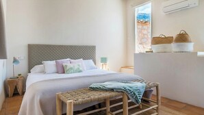 A bedroom with a bed, bedside tables, a bench, a window with a view, and a shelf with baskets. - Design stone house in Mallorca's Sóller valley (Soller)