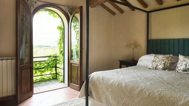 Un dormitorio con una cama grande, una mesita de noche, una lámpara y una puerta abierta que da a un balcón con vistas a zonas verdes.