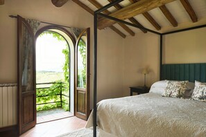 A bedroom with a large bed, a nightstand, a lamp, and an open door leading to a balcony with a view of greenery. - Restored barn in the Tuscan hills (Pergine Valdarno)