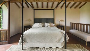 Una cama con dosel, con una colcha blanca, dos mesitas de noche con lámparas, y un banco a lo largo de la pared.