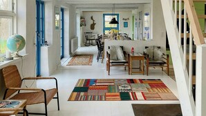 Een ruime loft met een hoge plafond, zichtbare houten balken, een eetgedeelte met een kleurrijk tapijt en een trap naar een hoger niveau.