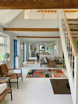Ein geräumiges Loft mit hoher Decke, freiliegenden Holzbalken, einem Essbereich mit farbenfrohem Teppich und einer Treppe zu einer oberen Ebene.