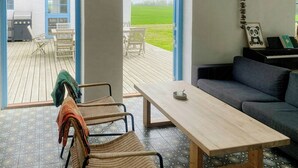 Een kamer met een houten tafel, een bank, rieten stoelen, een piano en uitzicht op een groen veld door open deuren.
