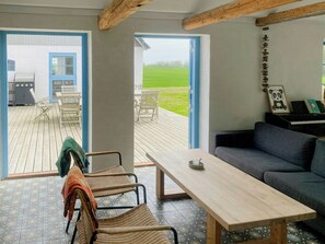 Ein Zimmer mit einem Holztisch, einer Couch, Korbstühlen, einem Klavier und Blick auf ein grünes Feld durch offene Türen.