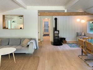 A modern living room with a sofa, a dining table, a black wood-burning stove, and a window with a view. - Renovated holiday home near Als Odde Beach (Hadsund)