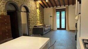 Une pièce aux murs de pierre, comprenant un canapé, une table et un coin cuisine avec un fourneau.
