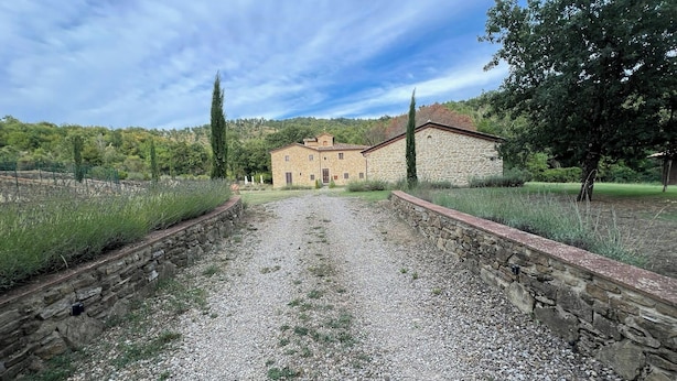 Un bâtiment en pierre, avec un chemin de gravier menant à l’entrée, entouré de verdure et d’arbres.
