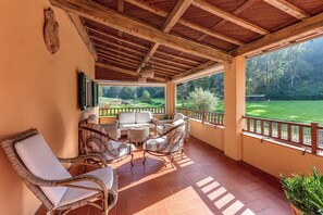 Eine überdachte Terrasse mit Korbmöbeln, Blick auf einen Golfplatz und Holzboden.