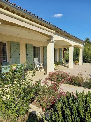 Ein überdachter Außenbereich mit Stühlen und Tischen, ein Garten mit verschiedenen Pflanzen und Blumen sowie ein Gebäude mit Ziegeldach und grünen Fensterläden.