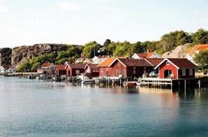 A waterfront property with red buildings, docked boats, and a rocky hillside in the background. - Historic timber cottage from 1700s on Hamburgö (Hamburgsund)