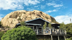 Un refugio de montaña con una gran formación rocosa al fondo, una terraza de madera y un cielo azul despejado.