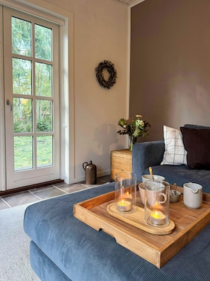 Ein Zimmer mit einem großen Fenster, einem Holfnachtkästchen, einem blauen Sofa und einem Tablett mit Kerzen.