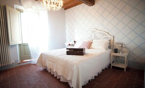Ein Schlafzimmer mit einem Bett, einer Kommode, einem Heizkörper, einem Fenster mit Vorhängen und einer Pendelleuchte.