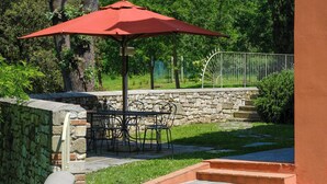 Eine Terrasse mit Tisch und Stühlen unter einem roten Sonnenschirm, umgeben von Steinmauern und Grünpflanzen.