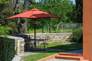 Eine Terrasse mit Tisch und Stühlen unter einem roten Sonnenschirm, umgeben von Steinmauern und Grünpflanzen.